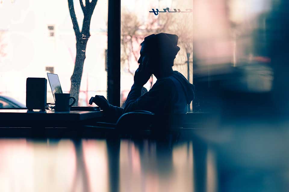 Person sitting at a table in a café, using a laptop, with a coffee cup, facing a large window.