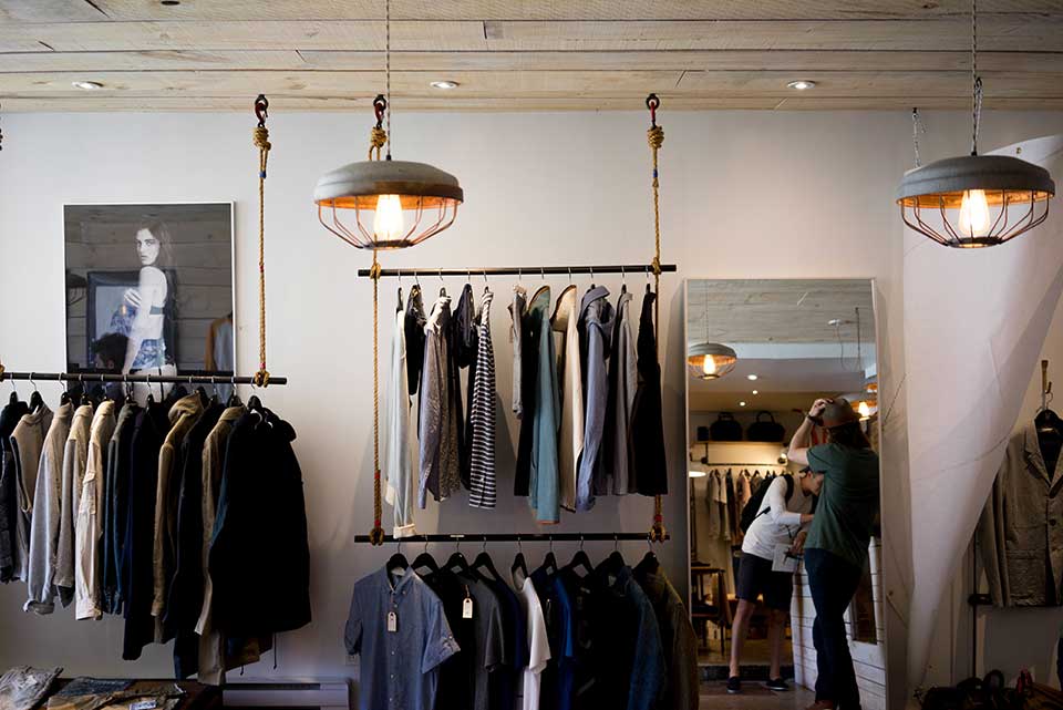 Clothing boutique with hanging lights, racks of clothes, and two people looking in a mirror at the back.