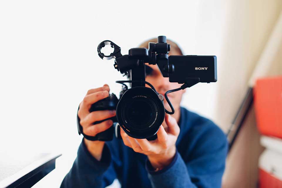 Person holding a Sony video camera, facing the camera lens toward the viewer in a brightly lit room.