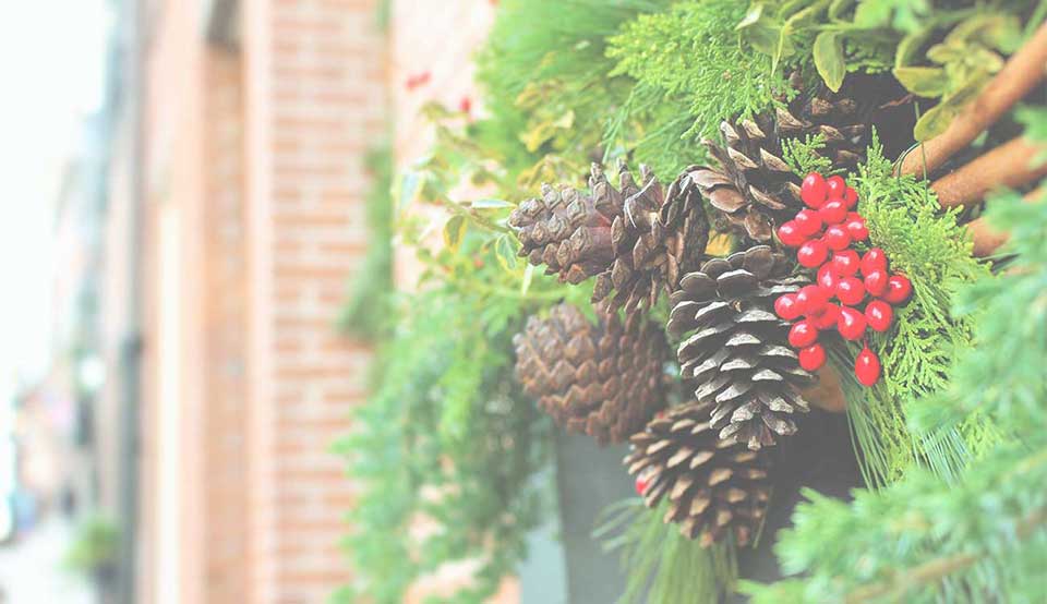 Festive wreath with pinecones, red berries, and green foliage hanging on a brick wall.