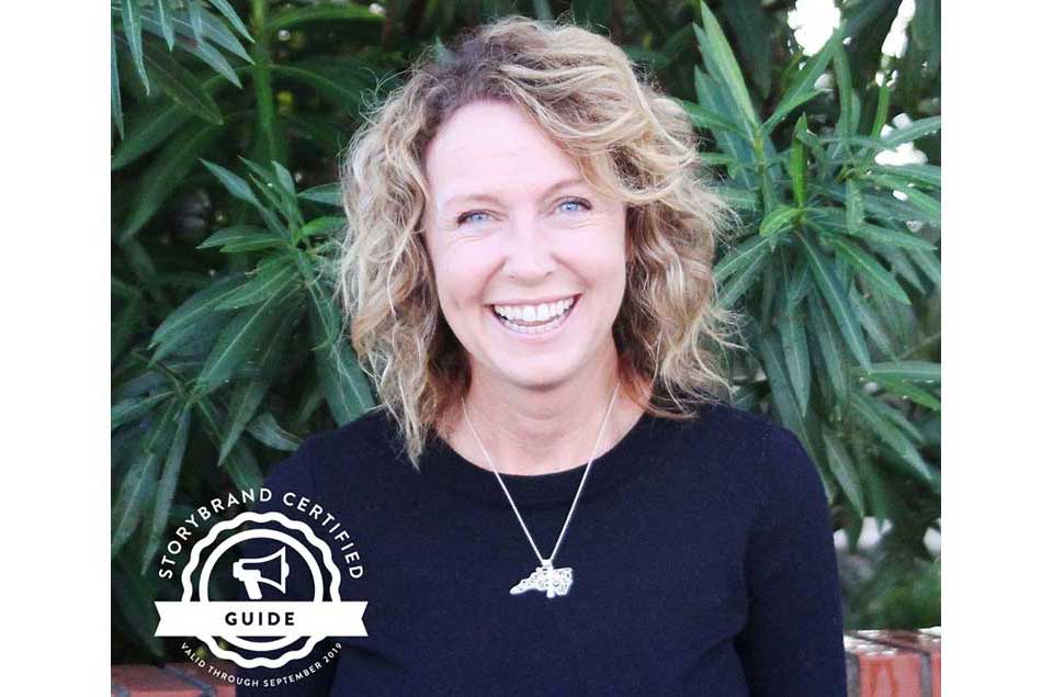 Smiling woman with curly hair wearing a black top, standing in front of green leafy plants.