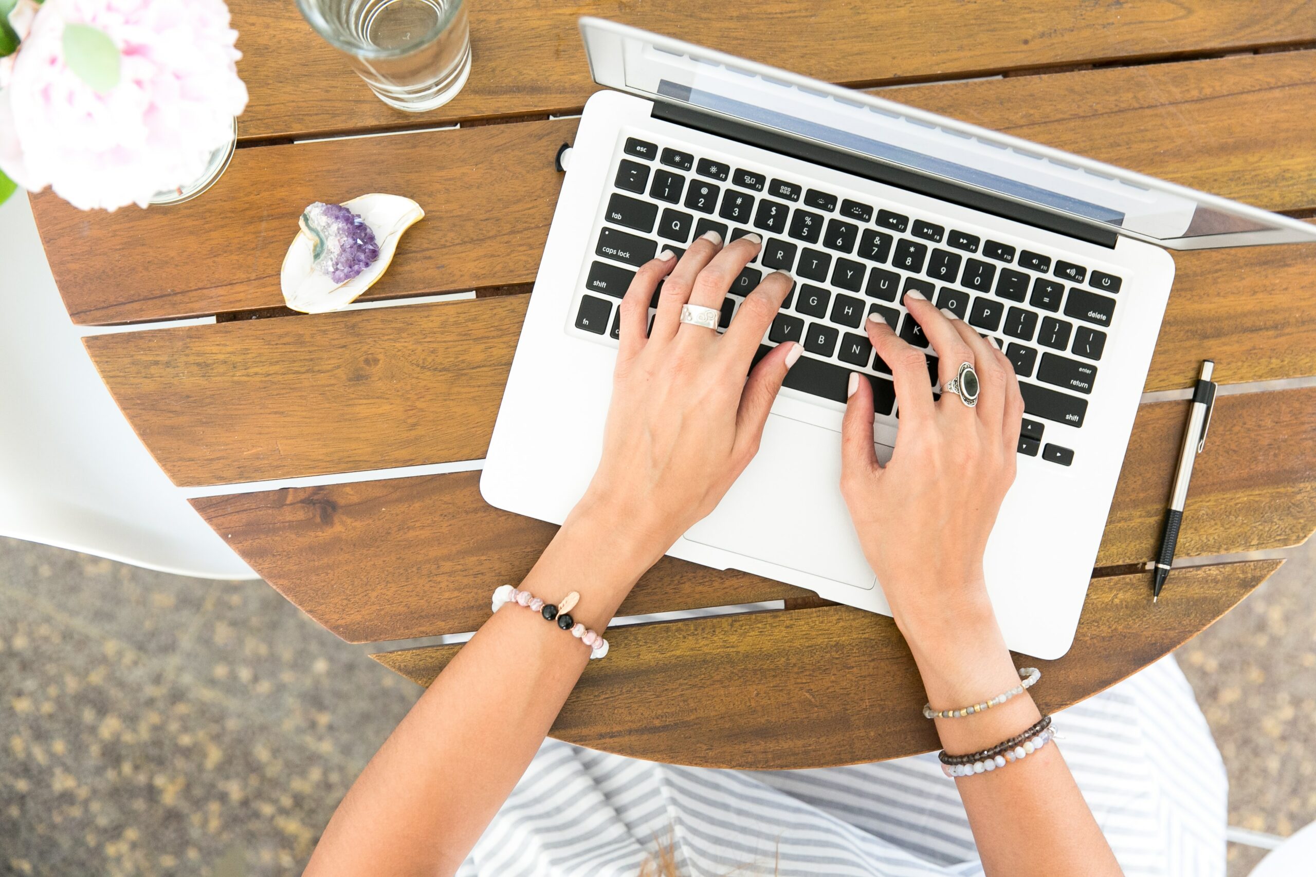 Hands typing on a laptop on a wooden table with jewelry, a pen, and a glass of water nearby.