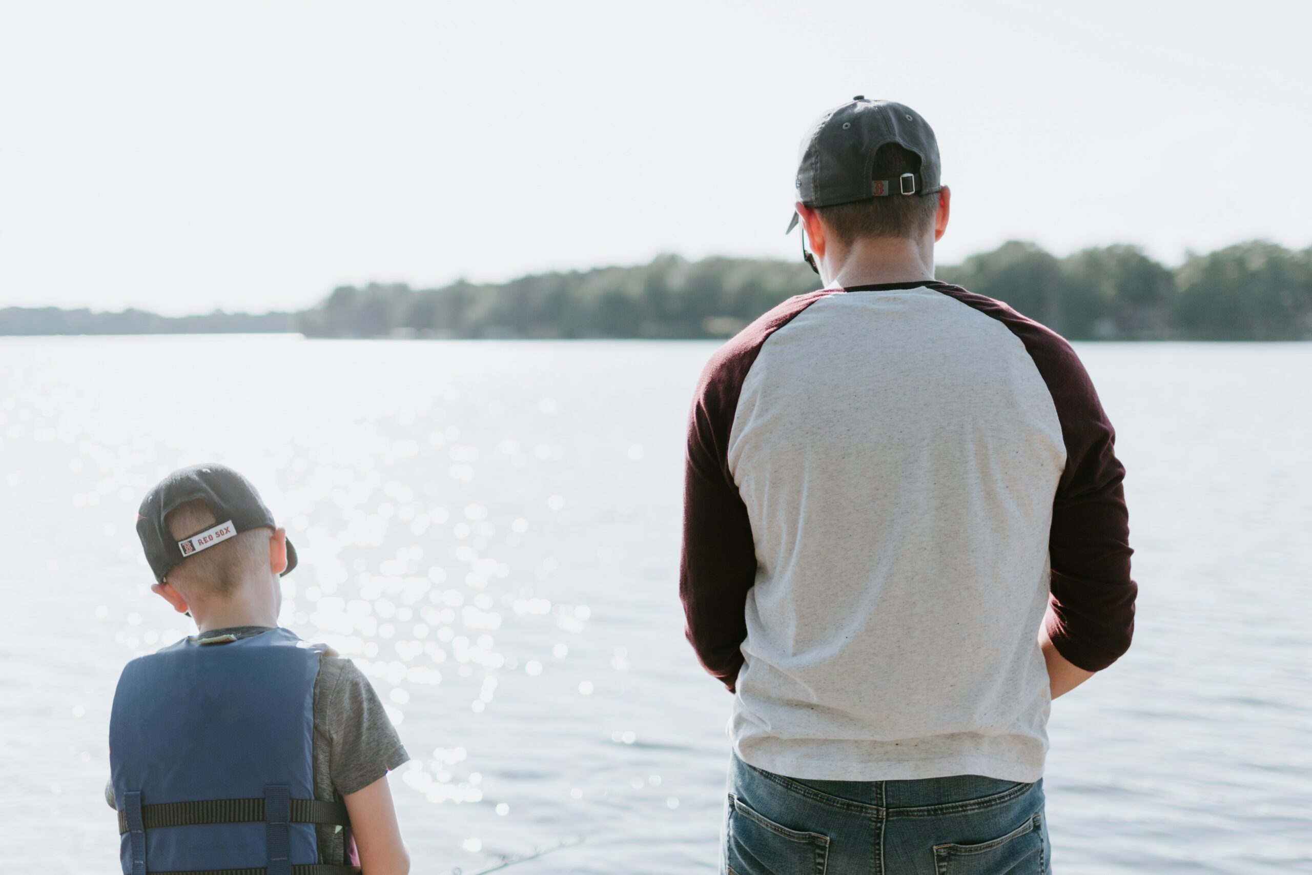 An adult and child wearing caps fish together by a sunlit lake, seen from behind.