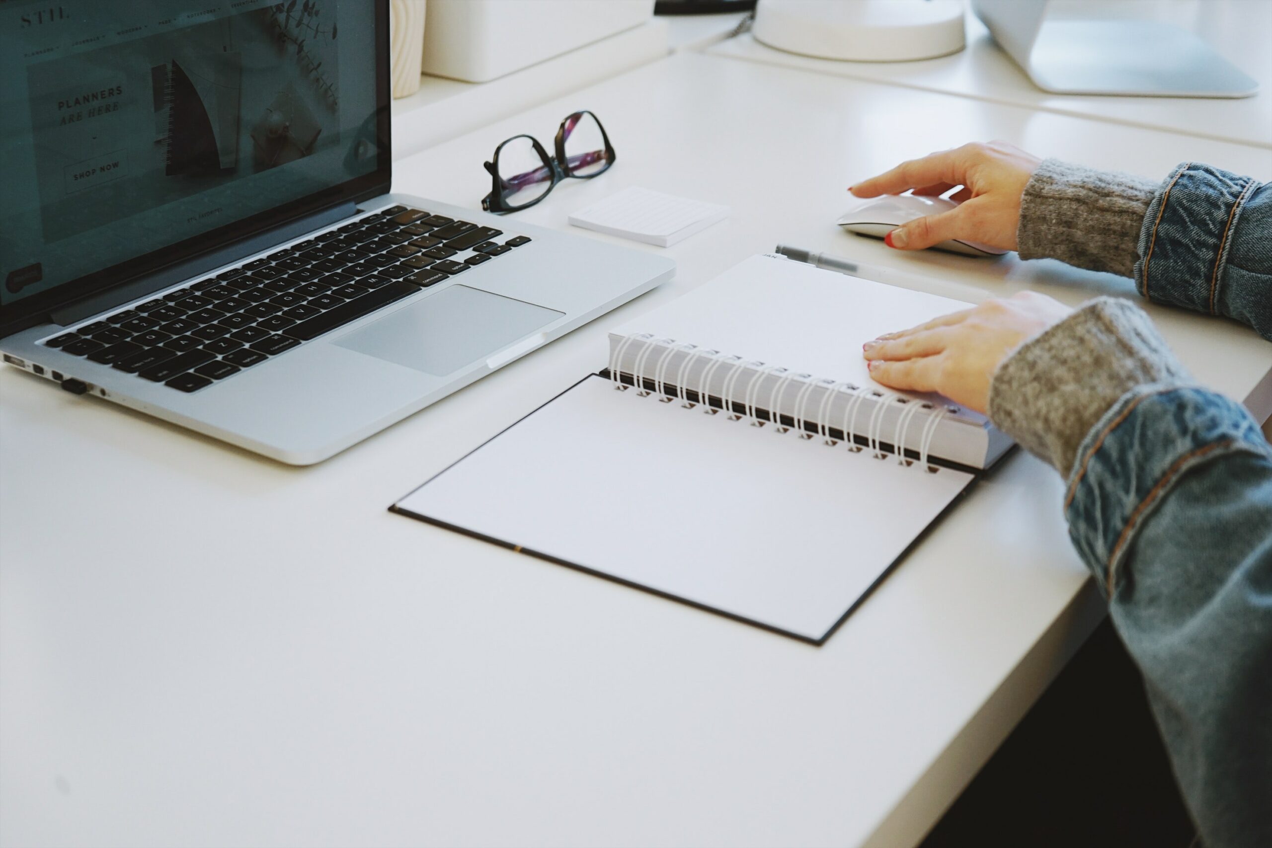 Person using a laptop and wireless mouse, with a blank notebook and glasses on a white desk.