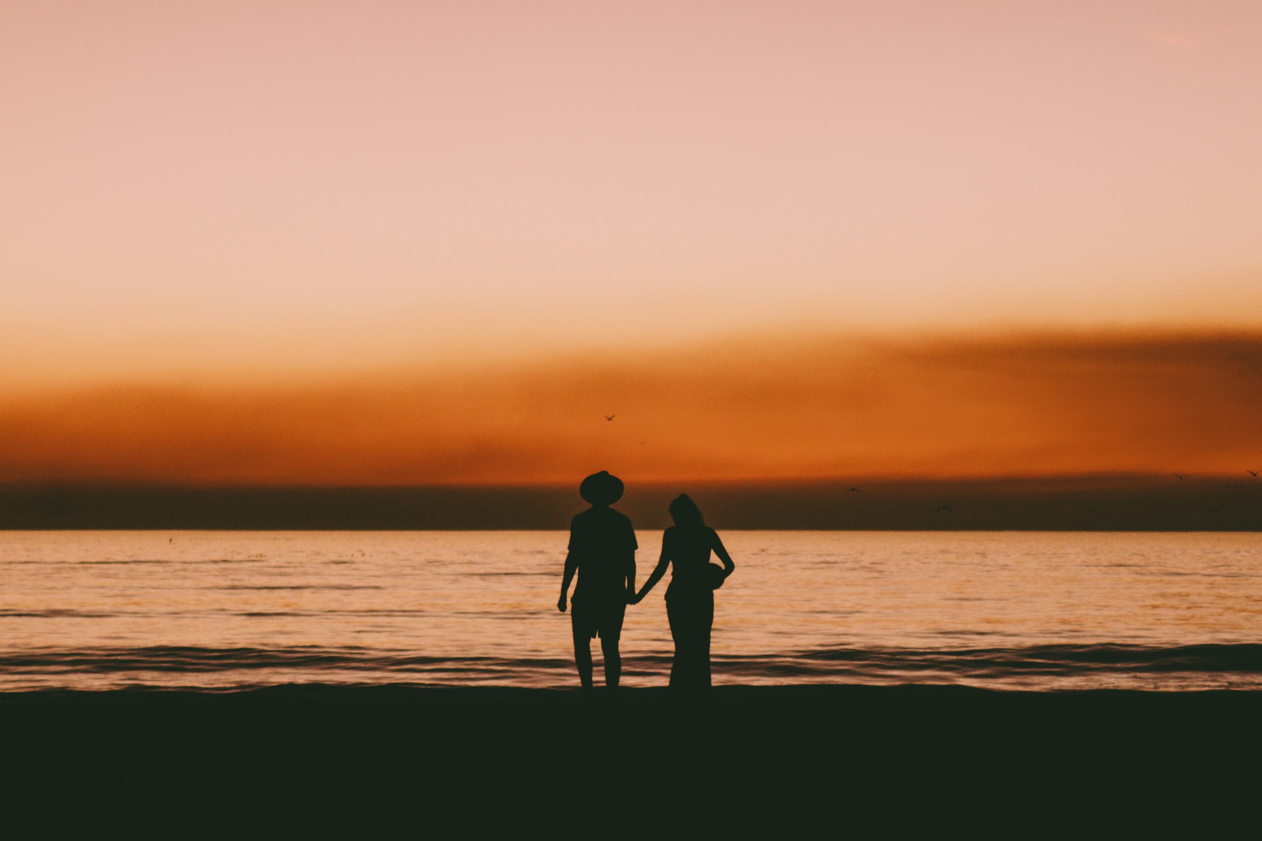 Silhouette of a couple holding hands on the beach at sunset with an orange sky in the background.