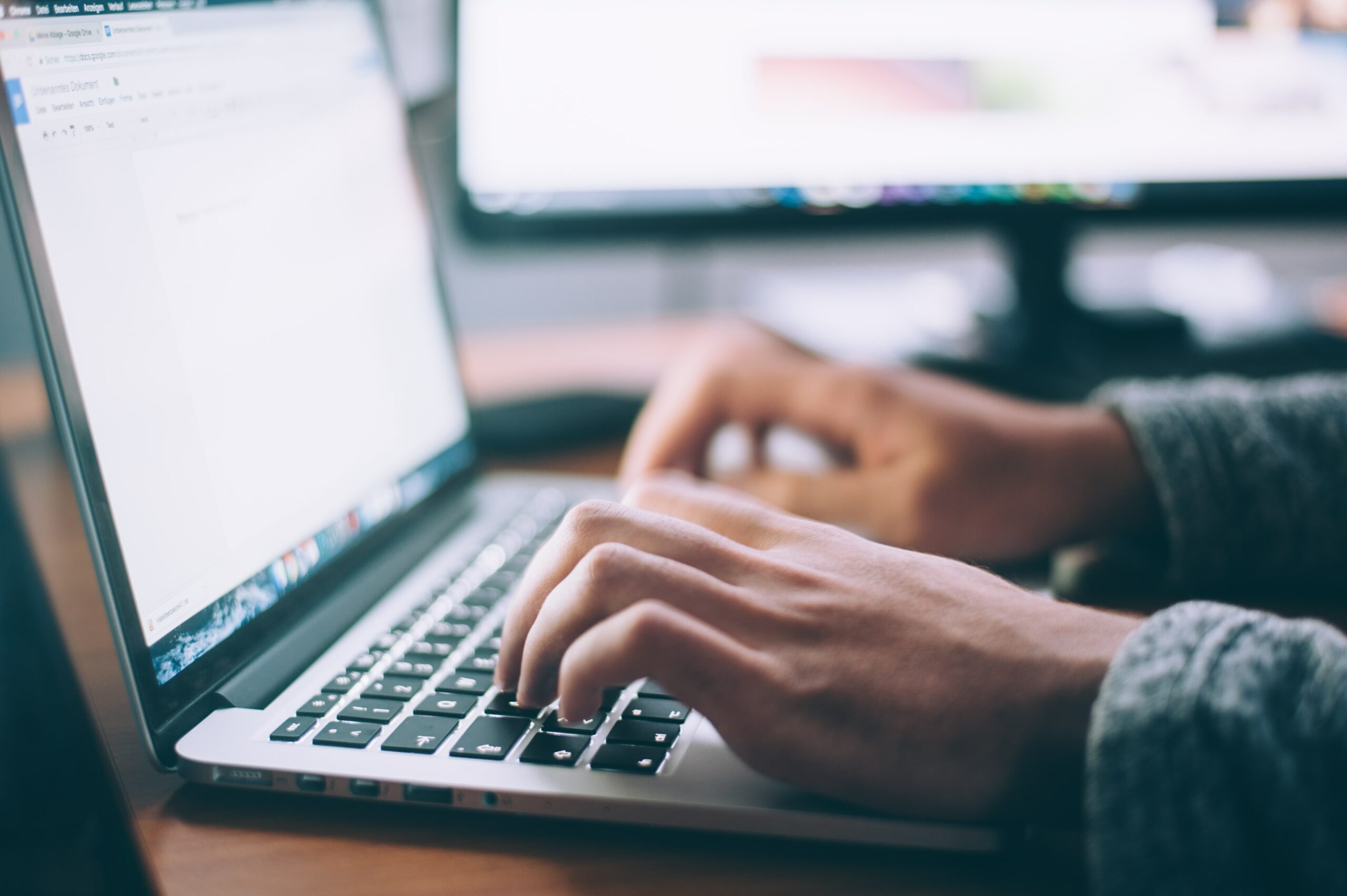 Hands typing on a laptop keyboard with two computer monitors in the background.