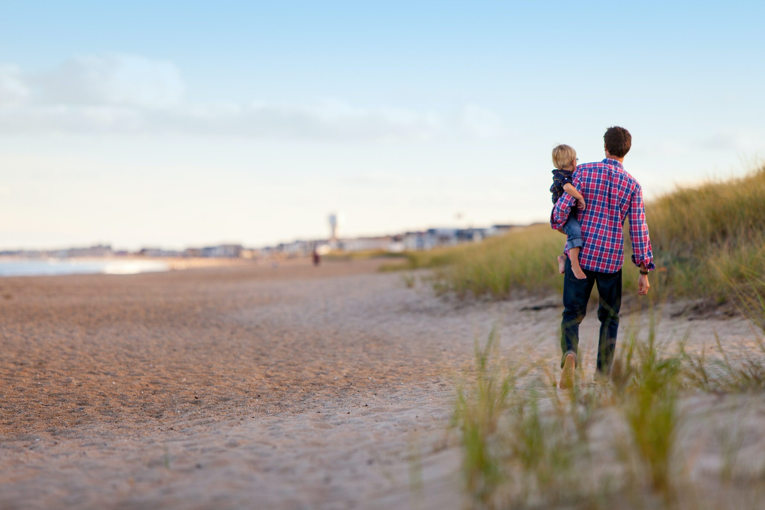 A man carries a child while walking along a sandy beach with grass and distant buildings.