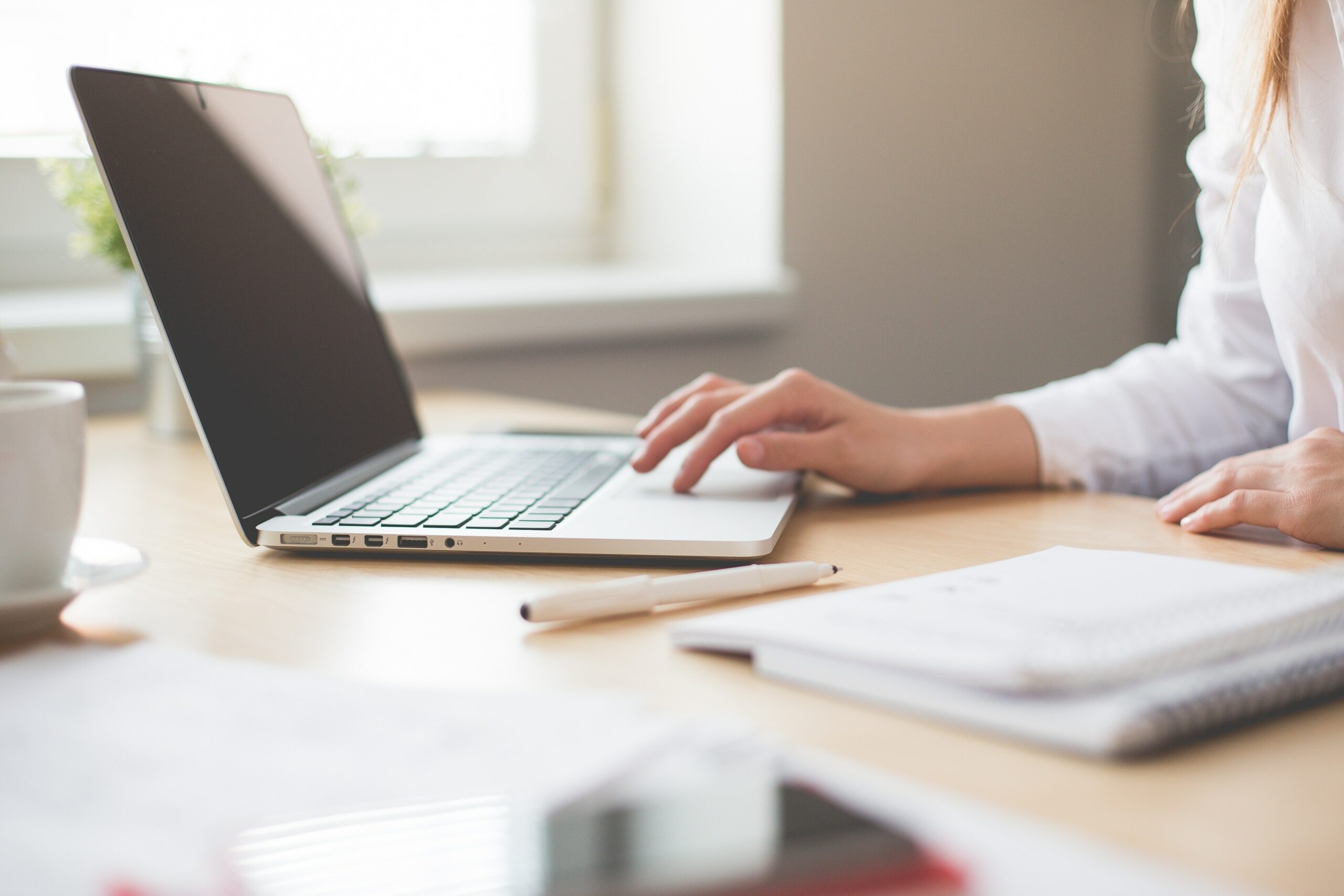 Person working on a laptop at a desk with notebooks, pen, and a window in the background.