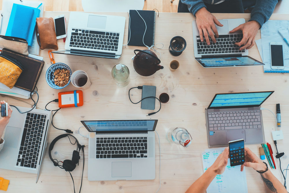 Four people working at a table with laptops, notebooks, snacks, and various tech accessories.