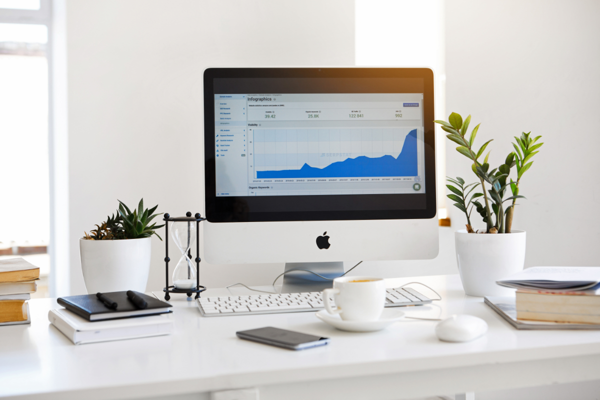 IMac displaying a blue analytics graph on a white desk with plants, books, a coffee cup, and office supplies.