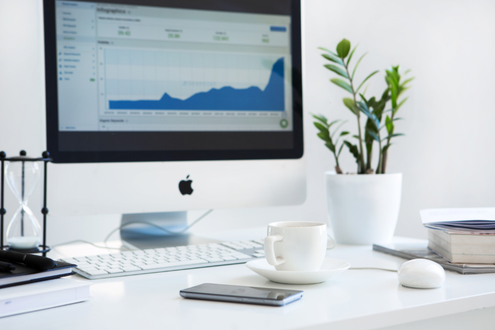 White desk with a computer displaying a graph, a coffee cup, phone, books, and a potted plant.