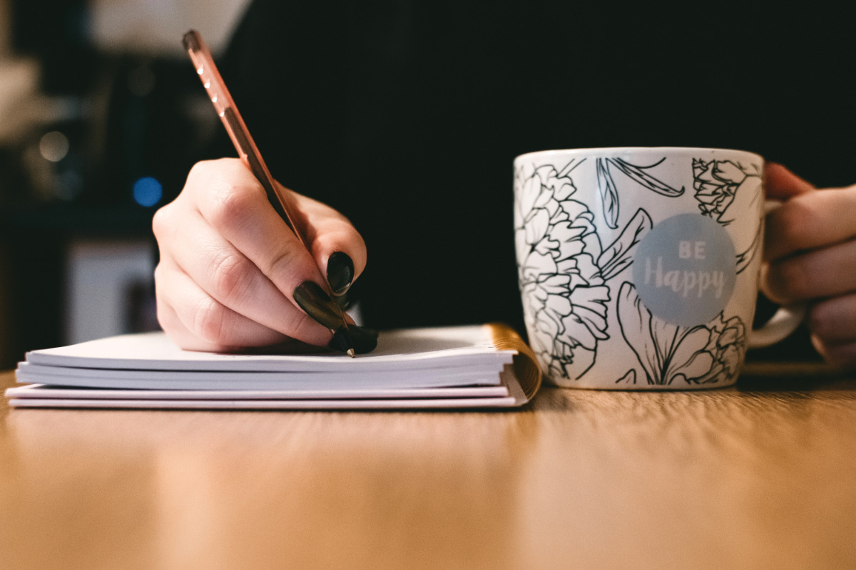 A person writes in a notebook while holding a floral mug that says "Be Happy" on a wooden table.