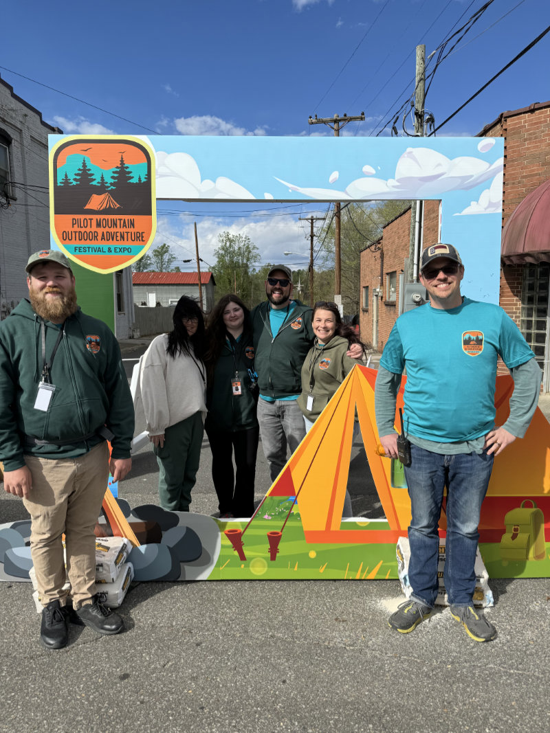 Six people pose outdoors in front of a colorful Pilot Mountain Outdoor Adventure photo frame display.