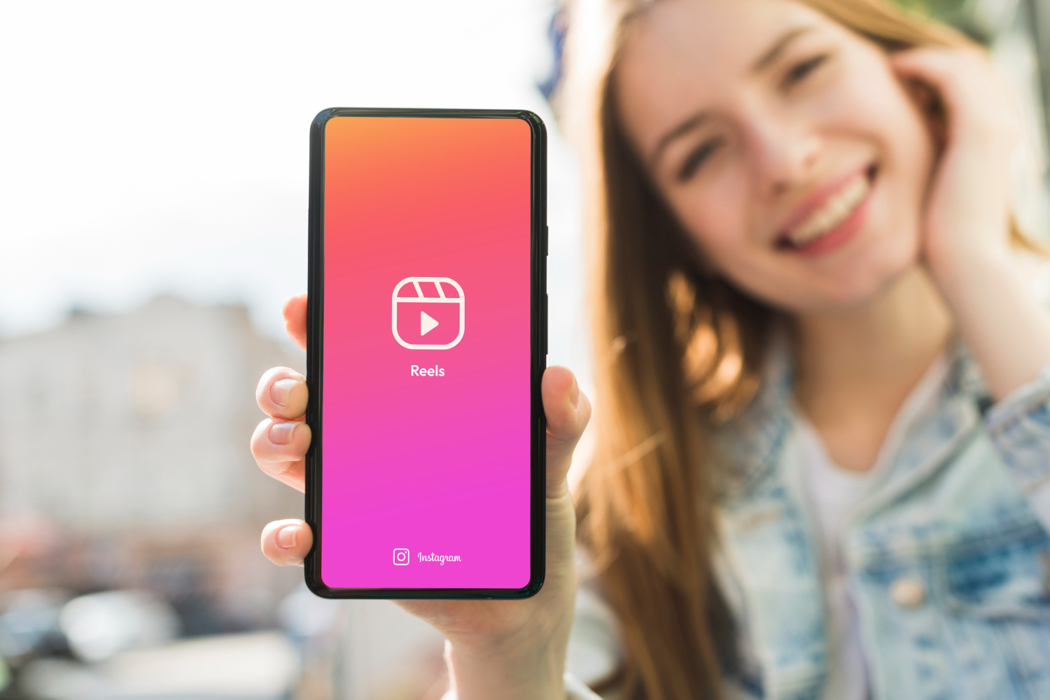 Smiling woman holds a smartphone showing the Instagram Reels logo on the screen, highlighting Instagram Reels as a powerful tool for SMB marketing.