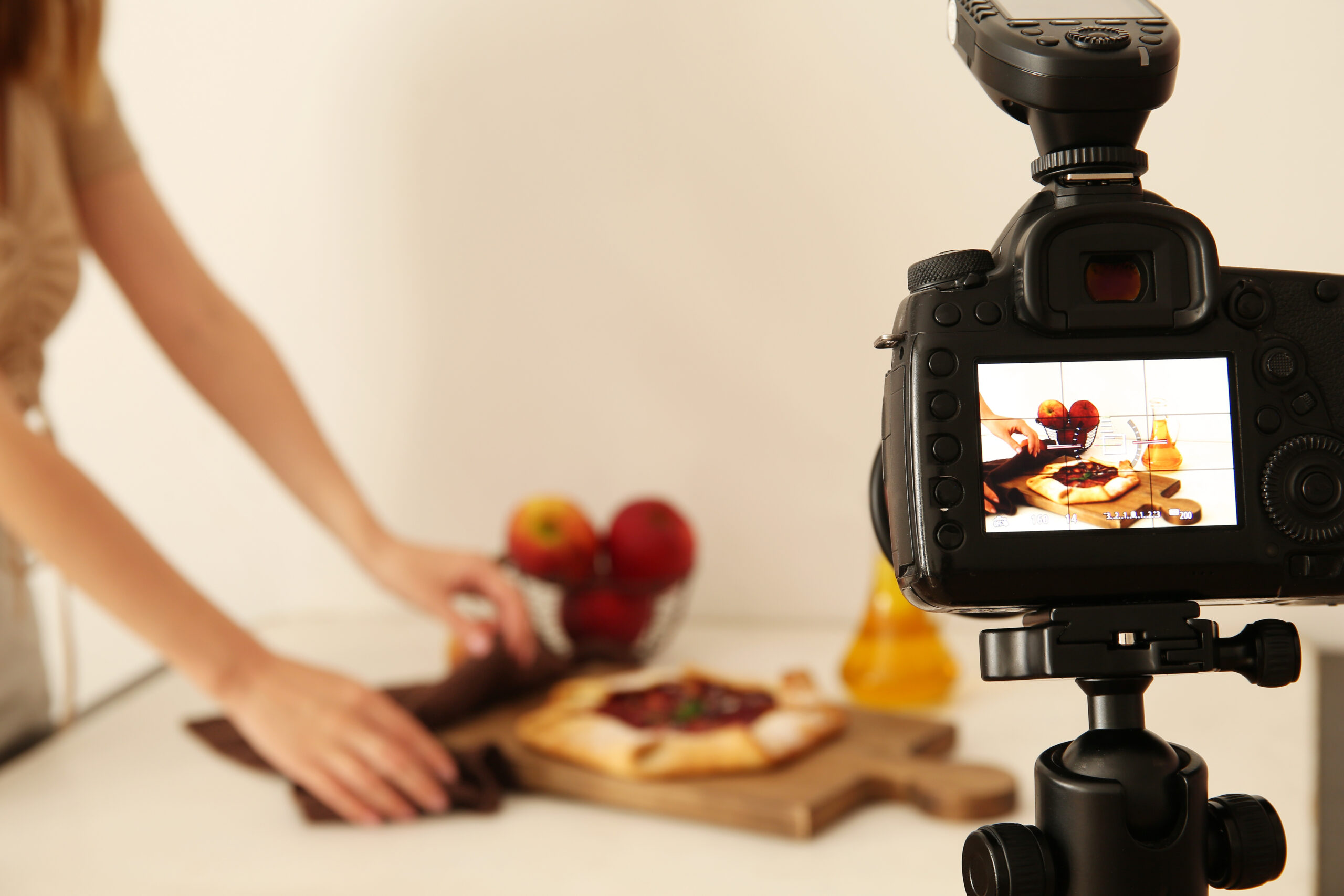 A camera films a person arranging food and fruit on a table with a cutting board and a bottle of oil, capturing the art of food photography in action.
