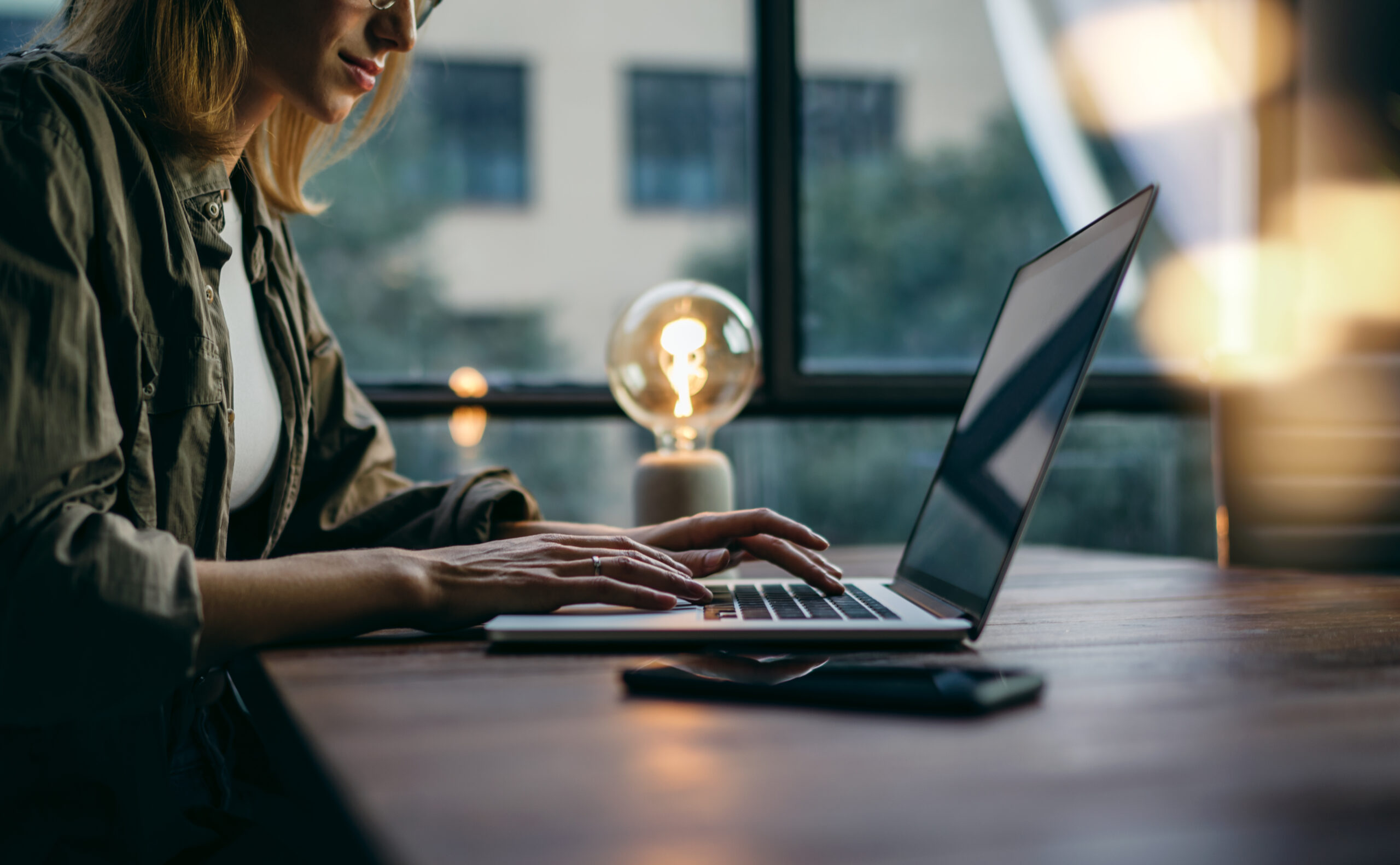 Person typing on a laptop at a wooden desk near a window, crafting a blog post with SEO in mind, with a lamp and phone in the background.