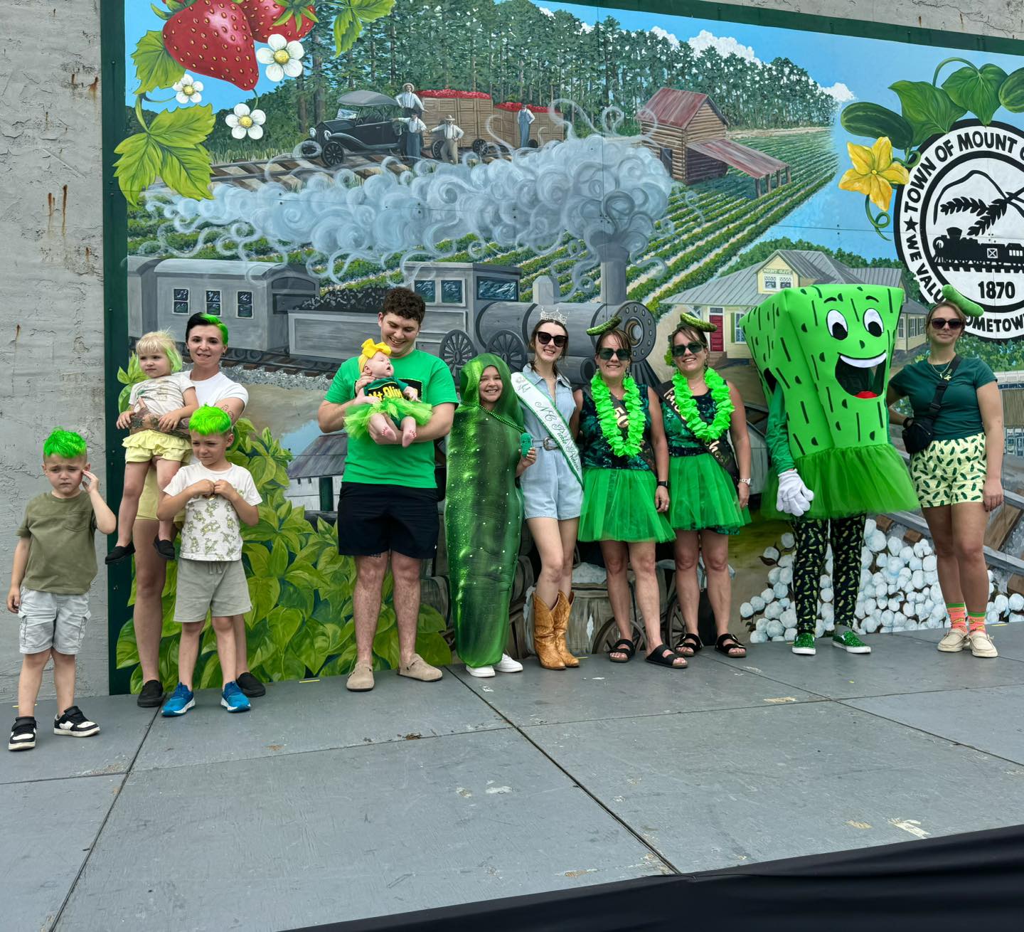A group of people dressed in green pose in front of a colorful mural with a cartoon pickle mascot, celebrating at a lively festival.