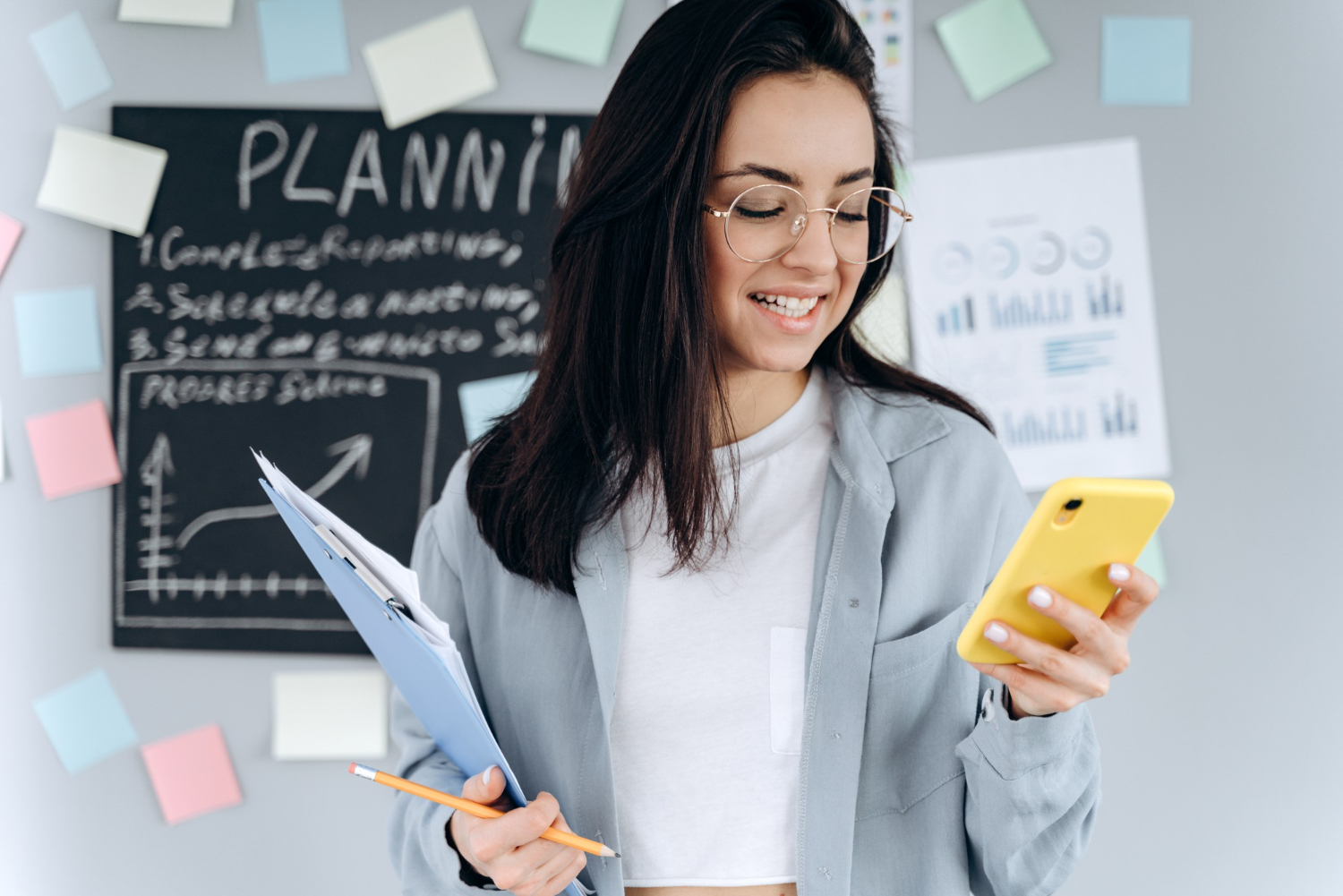 Smiling woman holding a clipboard and phone, standing by a chalkboard and colorful sticky notes.
